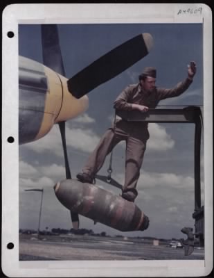 ␀ > Cpl. Lloyd Shumway Directs Crane With A 500 Lb. Bomb Into Place On The Wing Of A North American P-51 At A Base In England.