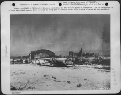 Thumbnail for Consolidated > Biscari airfield is littered withwrecks, including the two-motored plane in foreground. In the background is a large demolished hangar, from 5-9 July 49 heavy and 212 medium bomber sorties were directed at this airfield.